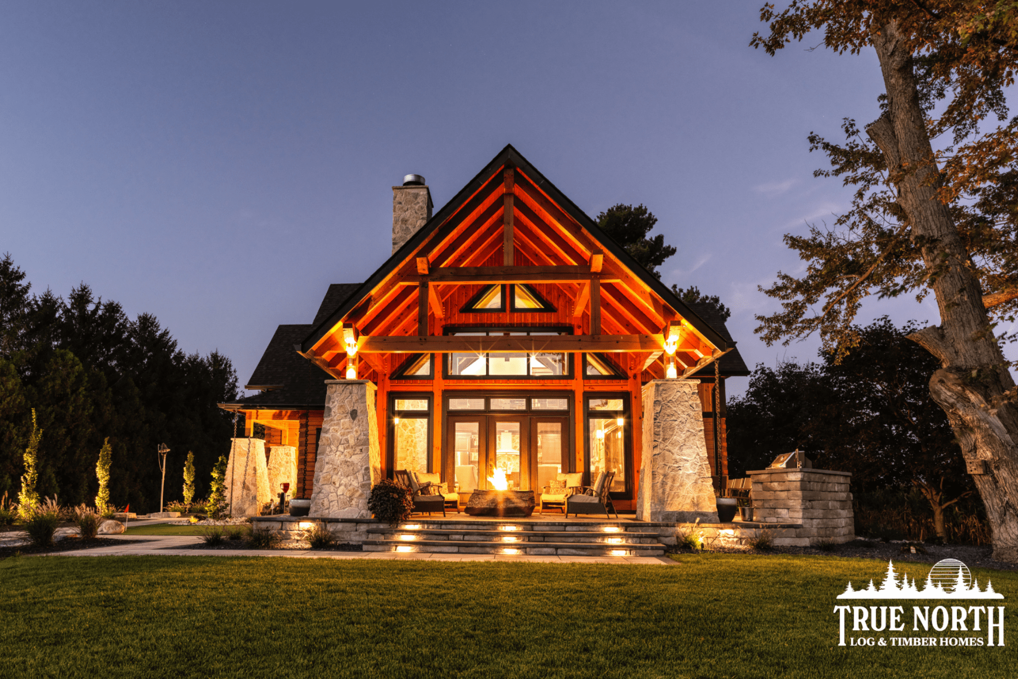 A log home with timber framing, taken at dusk. THis home was built by TrueNorth Log & Timber homes and is illuminated by the glow of a stone fireplace.